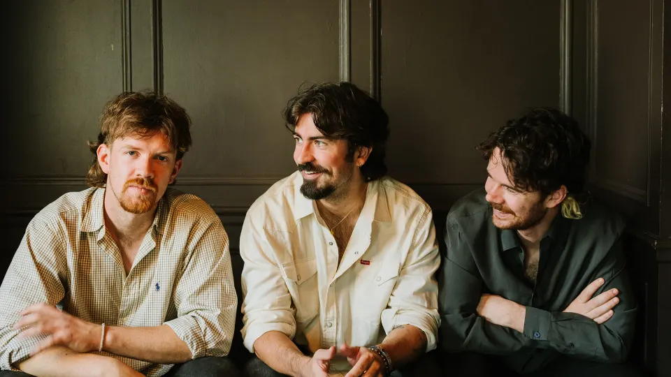 The three members of Amble sitting side by side against a dark panelled wall