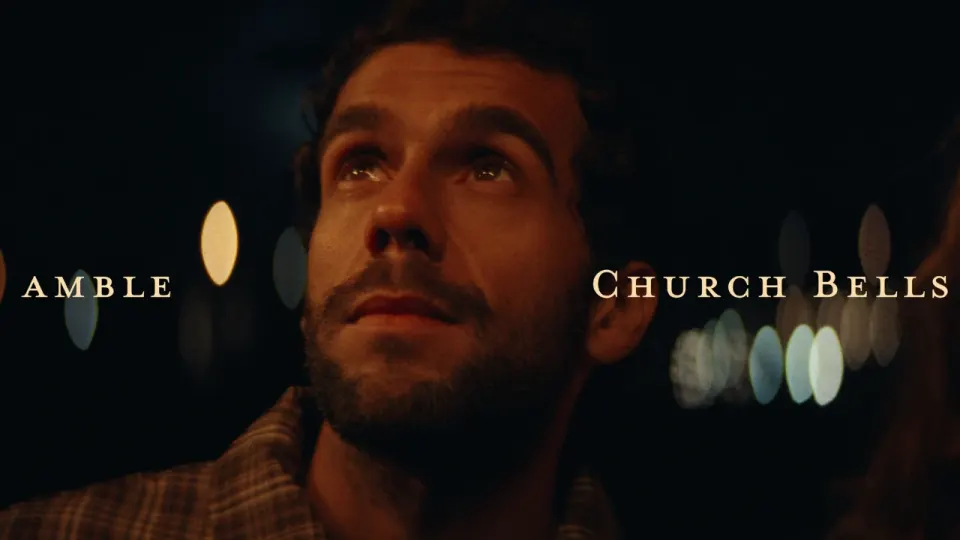 A man in a plaid shirt gazing upward at night, bokeh lights behind him, still from the Church Bells music video